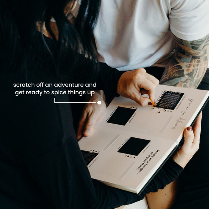"A couple and a hand scratching off a challenge in the 'In Bed' adventure book, revealing a surprise activity for couples."