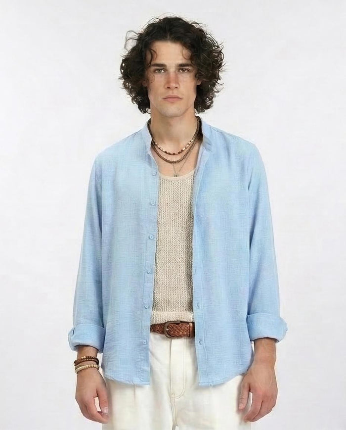 Man wearing a light blue shirt over a beige sweater with white pants on a plain background
