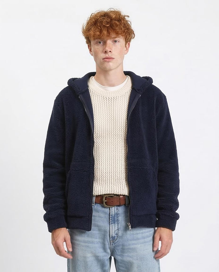 Man wearing a navy blue hoodie over a cream sweater with light blue jeans on a white background