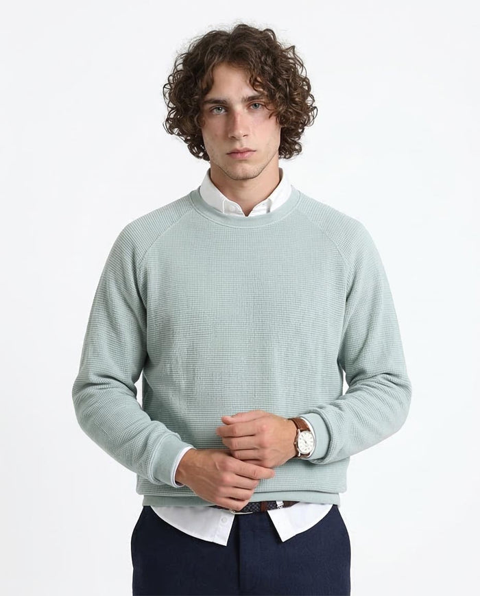 Man wearing a light green sweater over a white shirt with dark pants on a white background