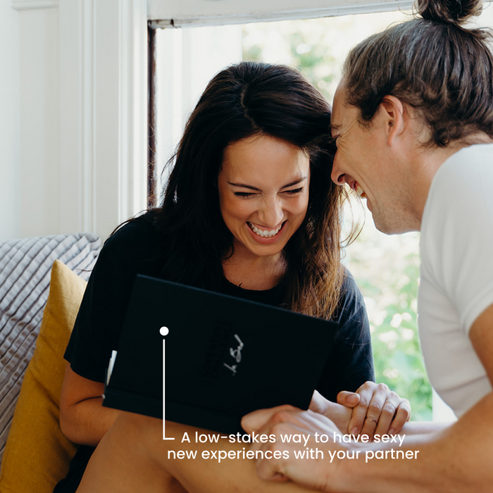 Couple laughing while flipping through the 'In Bed' challenge book, ready to pick their next activity.
